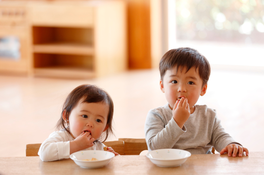 Two children sitting at a table eating from bowls in a bright room.